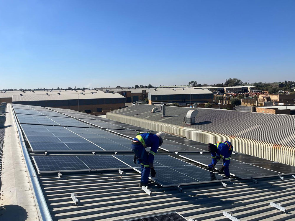 Two Quality Energy Solutions technicians installing solar panels on a commercial corrugated metal roof.
